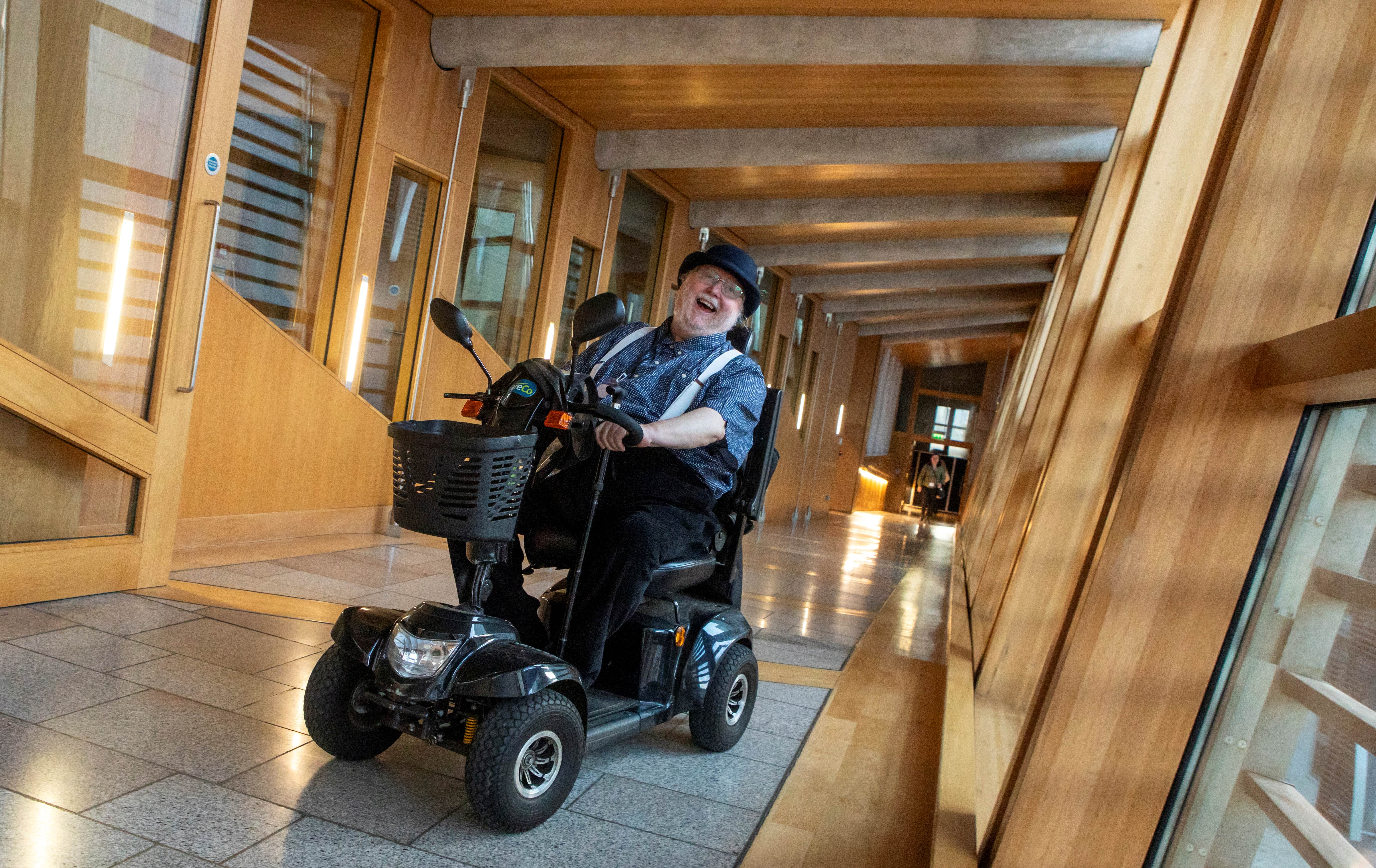 A smiling man wearing a hat moves through the corridors of the Scottish Parliament on his mobility scooter.
